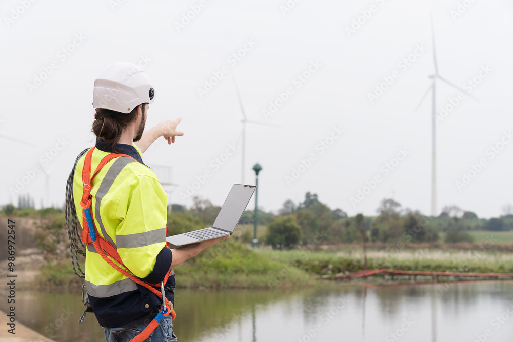 Male engineer working with laptop computer for maintenance of wind turbines at windmill field farm. Male engineer repairing or maintaining wind turbines at wind turbines farm Male engineer working with laptop computer for maintenance of wind turbines at windmill field farm. Male engineer repairing or maintaining wind turbines at wind turbines farm