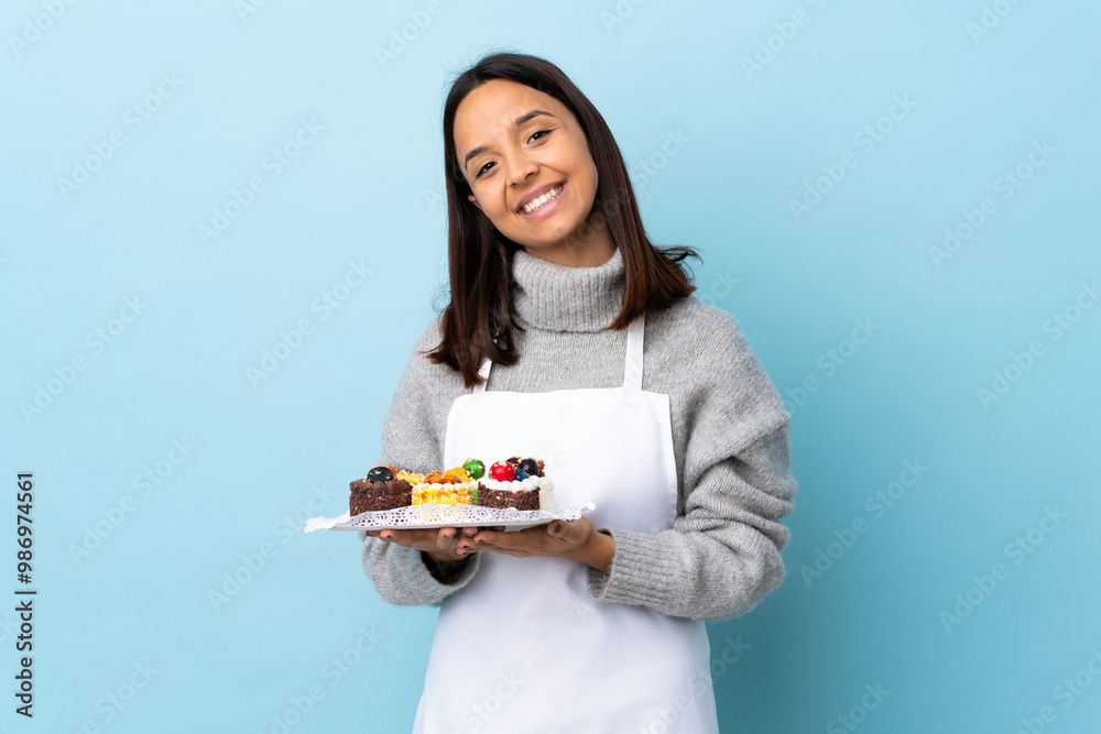 Pastry chef holding a big cake over isolated blue background keeps palm together. Person asks for something. Pastry chef holding a big cake over isolated blue background keeps palm together. Person asks for something.