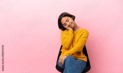Young mixed race woman sitting on a chair isolated on pink background making sleep gesture in dorable expression