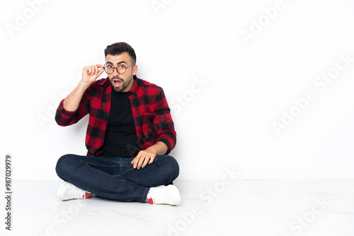 Young handsome man sitting on the floor with glasses and surprised