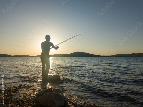 Fishing for pike, perch, carp. Fisherman with rod, spinning reel on sea or ocean. Man catching fish, pulling rod while fishing on sea, pond. Wild nature. The concept of rural getaway. Back view. photo