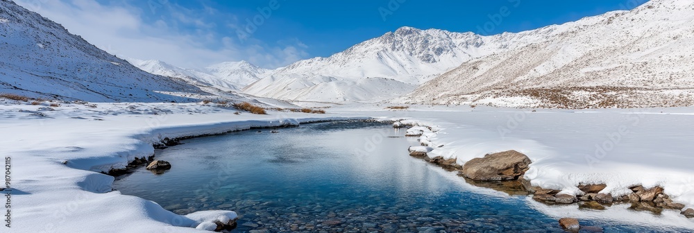 Fototapeta premium A crystal-clear blue sky backs snow-covered mountains and foreground rocks, encircled by flowing water