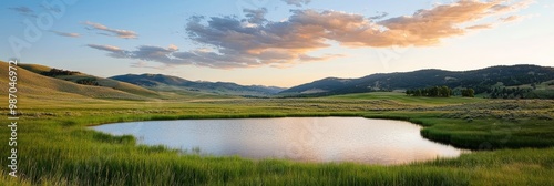  A tranquil pond in a field of green grass Mountains loom in the backdrop Clouds scatter the sky