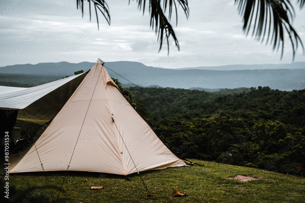 Obraz premium A triangle-shaped tent is pitched on a grassy hill, overlooking a lush green forest and distant mountains under a cloudy sky, creating a serene and scenic camping atmosphere.