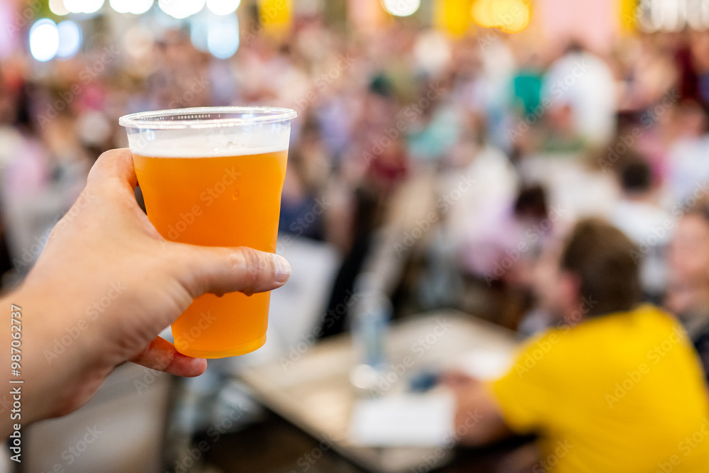 A close-up of a hand holding a plastic cup filled with amber-colored beer, with an out-of-focus crowd in the background. The setting suggests a social event or festival A close-up of a hand holding a plastic cup filled with amber-colored beer, with an out-of-focus crowd in the background. The setting suggests a social event or festival
