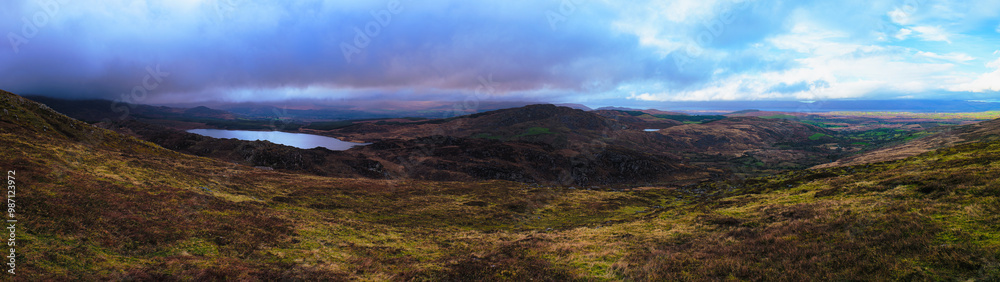 Fototapeta premium A panoramic landscape shot of rolling hills and a lake, with a cloudy sky above. The foreground shows rocky terrain covered in short grass and shrubs. The lake sits in a valley, surrounded by hills.
