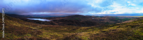 A panoramic landscape shot of rolling hills and a lake, with a cloudy sky above. The foreground shows rocky terrain covered in short grass and shrubs. The lake sits in a valley, surrounded by hills.