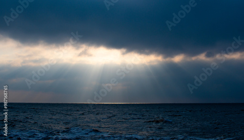 Sunrise over a dark sea with sunbeams breaking trough the clouds. A ribbon of sunlight stretches across the horizon.