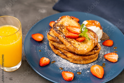 French toast garnished with strawberries and mint leaves with ice cream drizzled with caramel sauce and powdered sugar on a blue plate accompanied by a glass of orange juice on a dark background