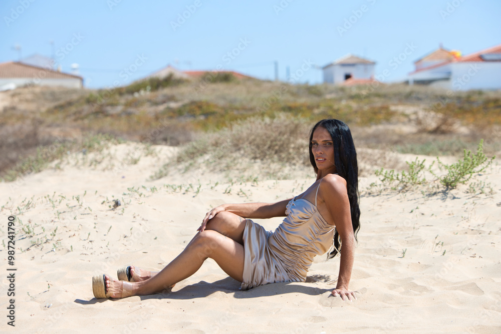 beautiful young brunette woman with long hair lying on a fine golden sand dune on a beach in the south of Spain in andalusia. The woman is wearing make-up, a long golden dress and high heels.