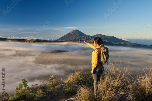 Asia, Indonesia, Java, Bromo Semeru national park, mature female tourist trekking at mount Bromo at sunrise, standing at viewpoint using mobile phone to take photographs

