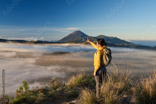Asia, Indonesia, Java, Bromo Semeru national park, mature female tourist trekking at mount Bromo at sunrise, standing at viewpoint using mobile phone to take photographs

