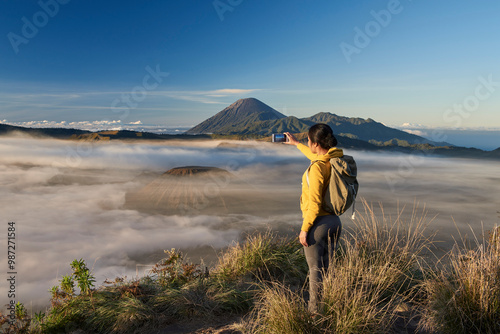 Asia, Indonesia, Java, Bromo Semeru national park, mature female tourist trekking at mount Bromo at sunrise, standing at viewpoint using mobile phone to take photographs

