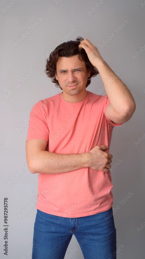 Confused man thinks scratching head by hand looking at camera on gray background. Curly middle aged guy feeling misunderstanding, trying to figure out situation. Pensive male wear jeans pink t-shirt. Confused man thinks scratching head by hand looking at camera on gray background. Curly middle aged guy feeling misunderstanding, trying to figure out situation. Pensive male wear jeans pink t-shirt.