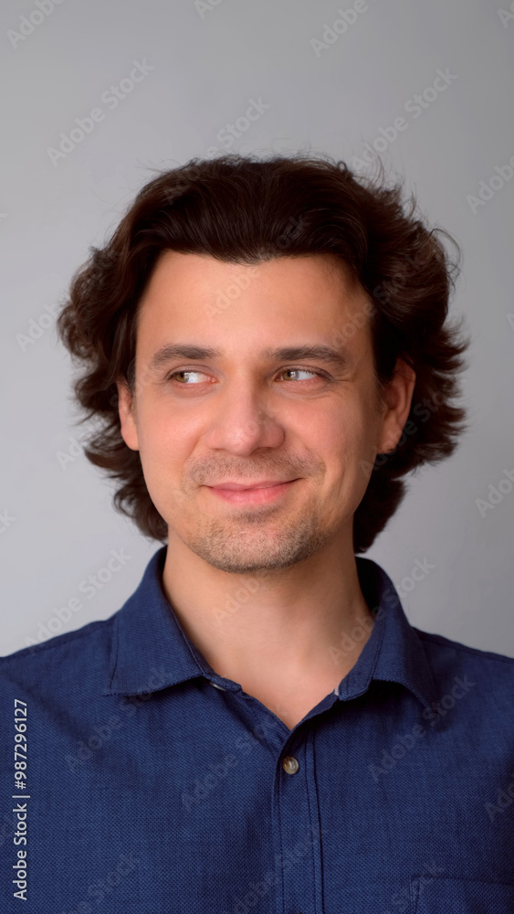 Portrait of man looking by side with sly tricky face on gray background. Curly middle aged guy having tricky plan in mind with smile. Wearing blue shirt. Showing emotions through facial expressions. Portrait of man looking by side with sly tricky face on gray background. Curly middle aged guy having tricky plan in mind with smile. Wearing blue shirt. Showing emotions through facial expressions.