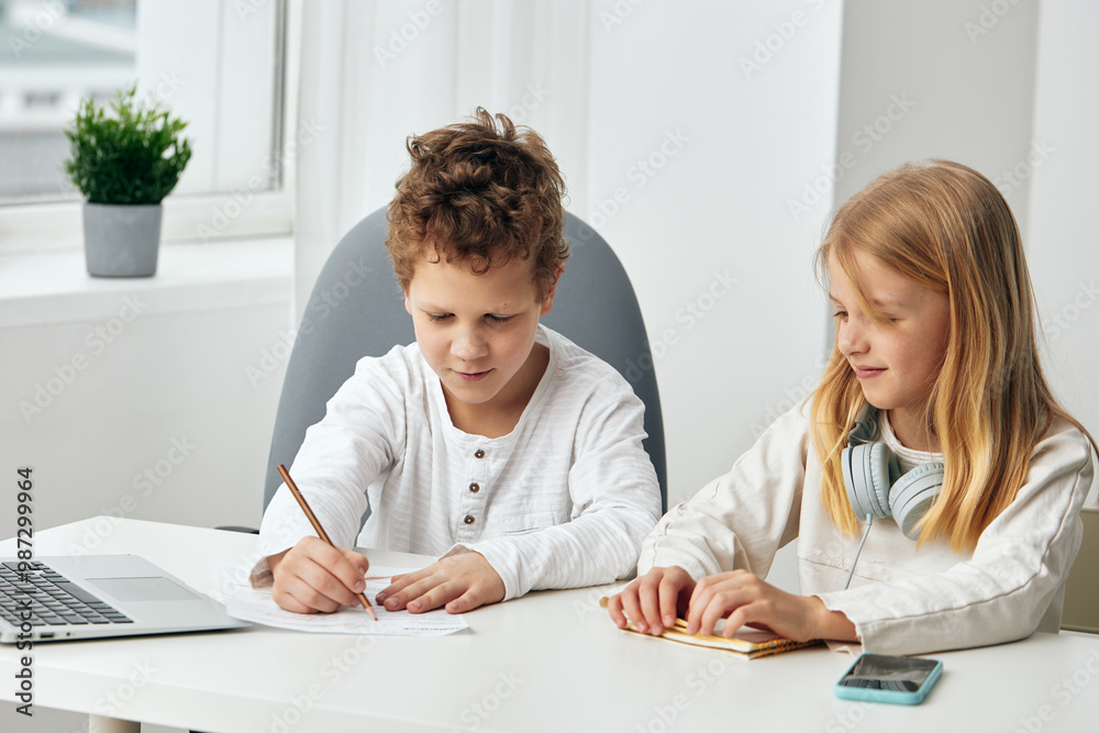 Happy children studying and playing online together at home a boy and a girl sitting at a table with laptops, headphones, and a tablet They are fully engaged in their educational activities, creating Happy children studying and playing online together at home a boy and a girl sitting at a table with laptops, headphones, and a tablet They are fully engaged in their educational activities, creating