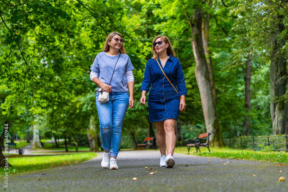 Fototapeta premium Two young beautiful smiling women dressed casual walking in city park on sunny day, trees in background. Meeting of friends. Walk in park. 