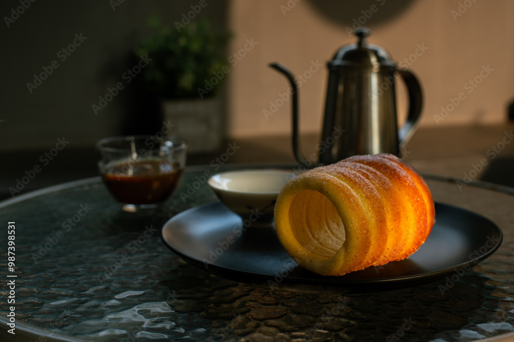Kurtoskalacs, hungarian sweet chimney cake on a black plate. Served as a breakfast alongside black coffee, in a cafe environment Kurtoskalacs, hungarian sweet chimney cake on a black plate. Served as a breakfast alongside black coffee, in a cafe environment