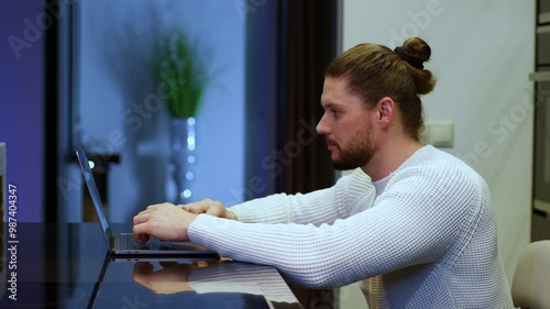 A man with a beard and a ponytail on his head works online from home, typing with his fingers on a computer. Freelancer work from home, online work from home. A man sits and solves problems.