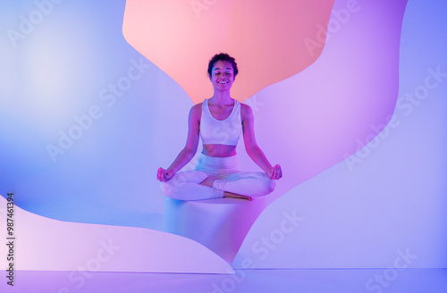 A young African woman in white fitness suit sits cross-legged in a meditation pose on round platform. Colored neon lights in blue and pink shades on background.