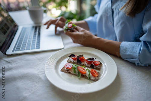 Tableau sur toile Unrecognizable female has rye crispbread sandwich at table, browsing online, working on laptop while eating