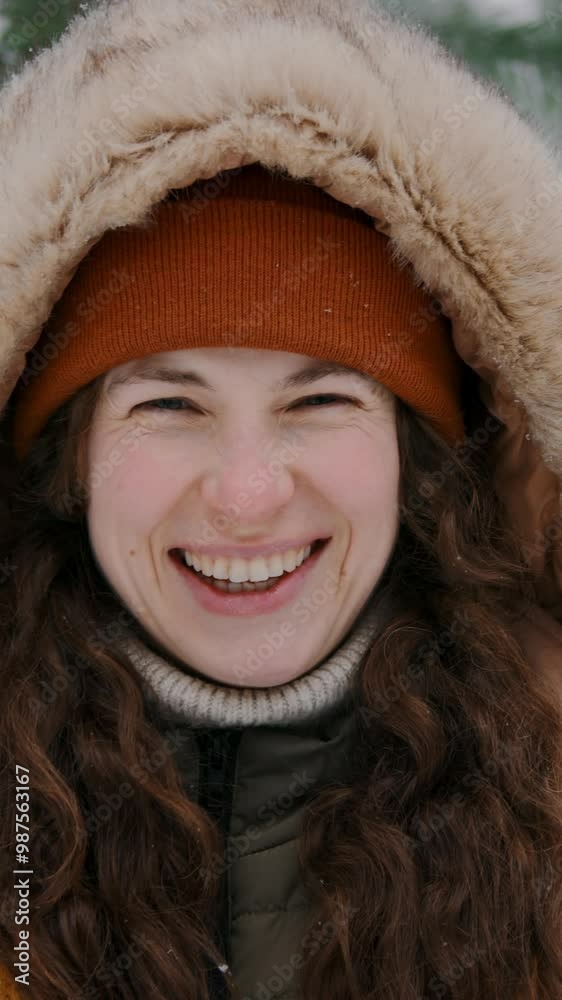 Close-up vertical portrait of cheerful young Caucasian woman in warm fur-trimmed hooded jacket, woolly knit hat standing in pine forest on winter day, looking at camera, laughing with joy Close-up vertical portrait of cheerful young Caucasian woman in warm fur-trimmed hooded jacket, woolly knit hat standing in pine forest on winter day, looking at camera, laughing with joy
