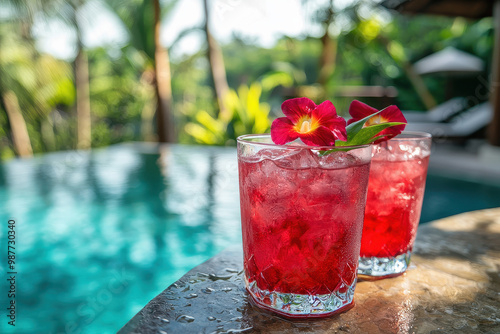 This image captures a beautiful scene with two refreshing red cocktails with ice and flowers, placed on a stone table by a luxurious poolside in a tropical paradise like setting. photo