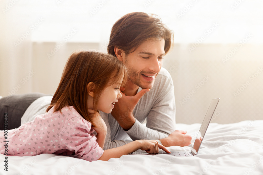 Little preschool girl learning how to use laptop, young father helping, lying on bed at home, free space Little preschool girl learning how to use laptop, young father helping, lying on bed at home, free space