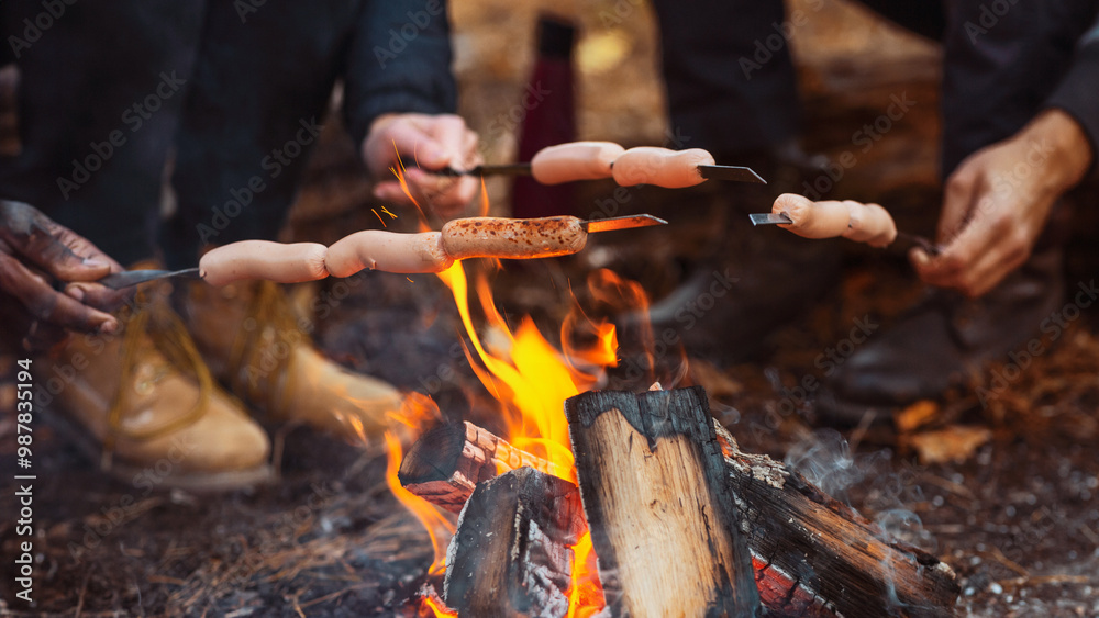 Fototapeta premium Cropped of mixed race group of friends sitting beside fireplace and frying sausages in forest