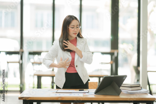 Overwhelmed and Stressed: A young Asian businesswoman clutches her chest, anxiety and exhaustion etched on her face as she battles work pressure in a modern office setting. 