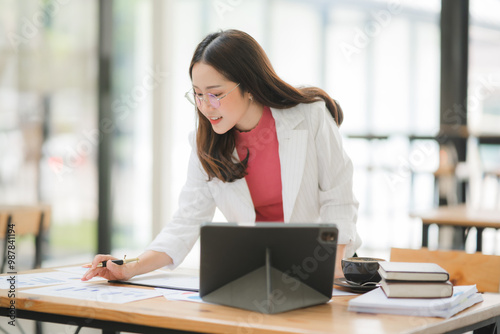 Focused Female Architect at Work: A young Asian woman architect engrossed in her blueprint designs, utilizing a digital tablet for modern architectural planning in a bright office setting. 