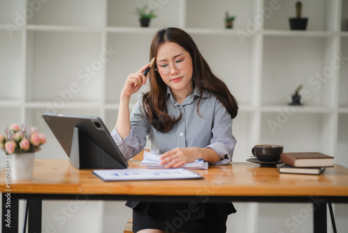 Businesswoman Concentrating: A focused young businesswoman in a modern workspace, absorbed in her work, with a thoughtful expression and a pen thoughtfully held to her temple, conveying a sense of ded photo