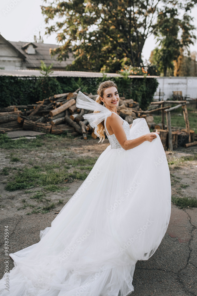 Fototapeta premium Beautiful young curly smiling blonde bride in a white dress outdoors dancing, spinning around herself against the background of logs in nature in the village.