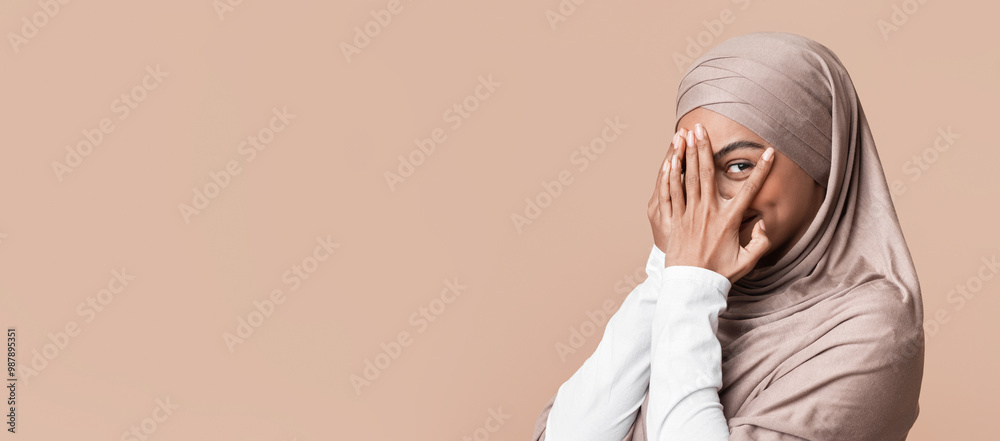 Closeup portrait of shy black muslim girl in headscarf peeking through fingers and looking at camera, yellow studio background with empty space Closeup portrait of shy black muslim girl in headscarf peeking through fingers and looking at camera, yellow studio background with empty space