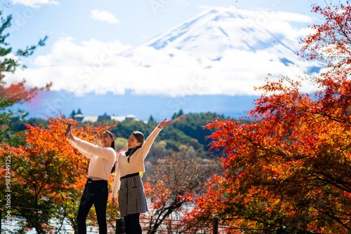 Happy Asian woman travel Japan Fuji on holiday vacation. Attractive girl friends using mobile phone taking selfie during travel Mt.Fuji and looking beautiful red maple tree leaf falling down in autumn photo