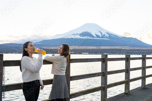 Happy Asian woman friend enjoy outdoor lifestyle travel nature in Japan on autumn holiday vacation. Attractive girl drinking tea from a bottle during travel Kawaguchi lake and Mount Fuji at sunset.