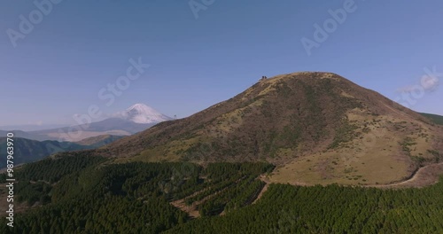 Aerial footage of a scenic mountain landscape with distant Mount Fuji in the background, featuring tree-lined hills and open grassy slopes, captured during a leftward tracking drone shot.