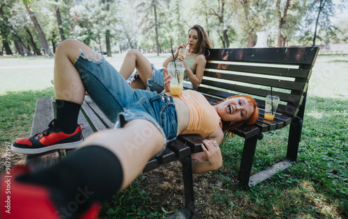 Two young adults relax on a park bench, enjoying a sunny day with refreshing drinks and playful smiles.