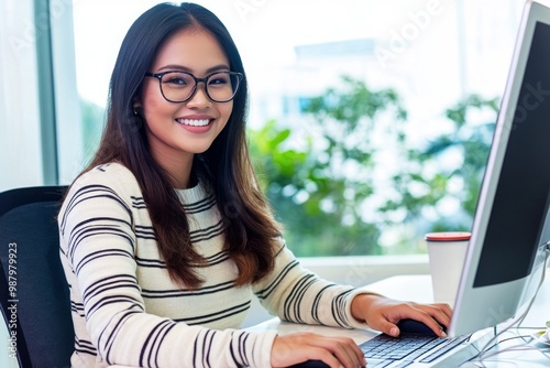 Smiling Filipino woman in glasses sitting at her desk, typing on a computer with a window view in the background.