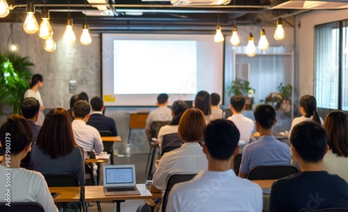 Asian business training presentation in a modern office, audience seated and listening to the speaker with light bulb pendant lights, whiteboard, and laptop displaying PowerPoint content.