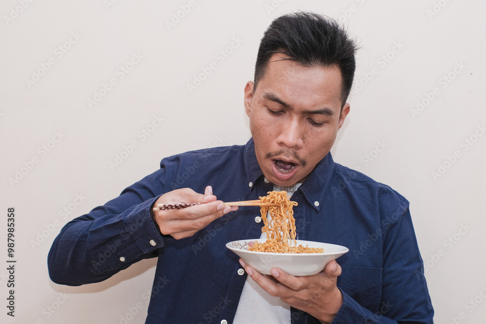 A Southeast Asian man wearing casual dress excited eat a plate of fried noodles and holding a plate of what instant noodles with chopsticks, the man expression is cheerful A Southeast Asian man wearing casual dress excited eat a plate of fried noodles and holding a plate of what instant noodles with chopsticks, the man expression is cheerful