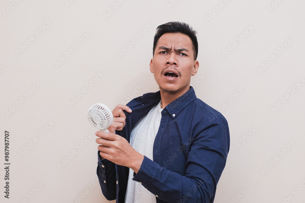 A Southeast Asian man wearing casual dress holding a small portable fan, his expression appears surprised or confused, as trying to cool himself down. A Southeast Asian man wearing casual dress holding a small portable fan, his expression appears surprised or confused, as trying to cool himself down.