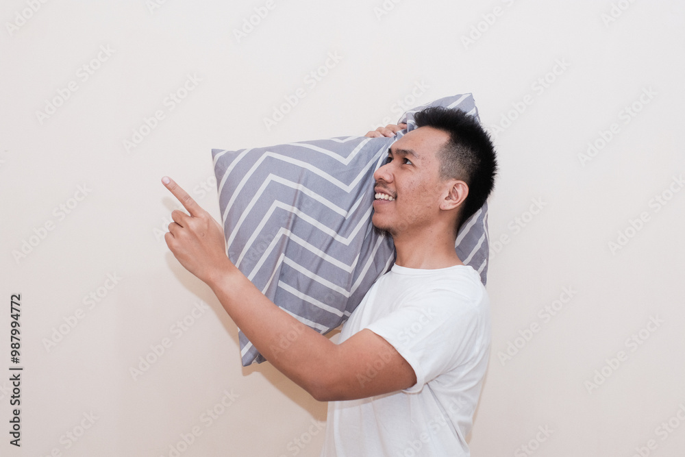A Southeast Asian man hold pillow pointing index finger aside up while resting at home isolated on white colour background studio portrait. Relax good mood lifestyle concept A Southeast Asian man hold pillow pointing index finger aside up while resting at home isolated on white colour background studio portrait. Relax good mood lifestyle concept