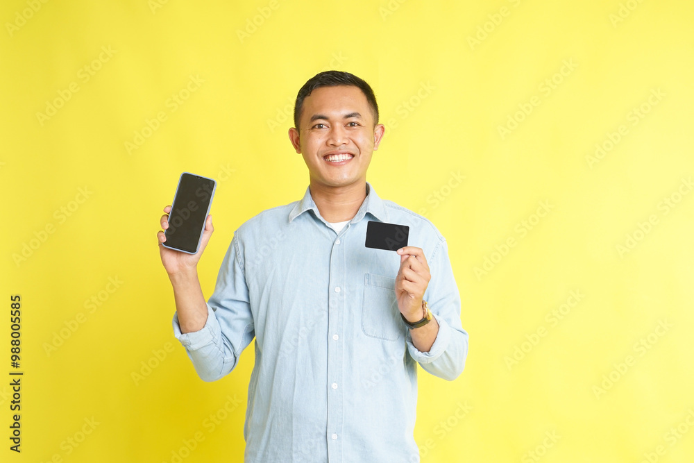Asian man in light blue denim shirt holding blank screen smartphone and blank card isolated on yellow background Asian man in light blue denim shirt holding blank screen smartphone and blank card isolated on yellow background
