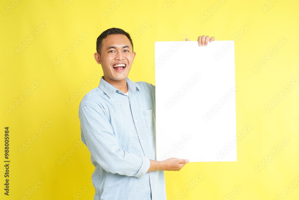 Asian man in light blue denim shirt holding blank white board for advertisement isolated on yellow background Asian man in light blue denim shirt holding blank white board for advertisement isolated on yellow background