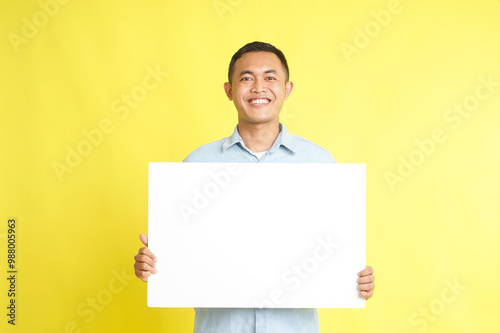 Asian man in light blue denim shirt holding blank white board for advertisement isolated on yellow background
