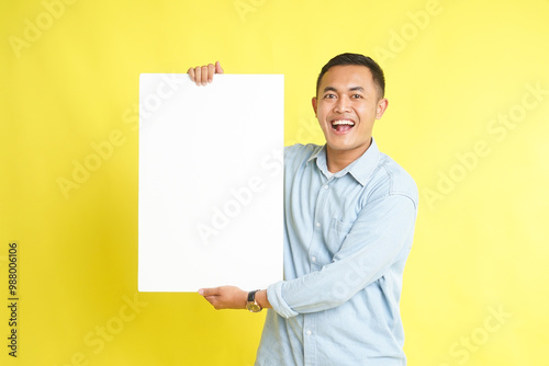 Asian man in light blue denim shirt holding blank white board for advertisement isolated on yellow background