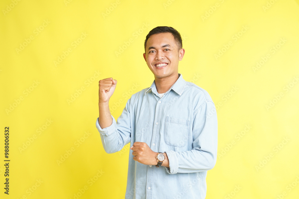 Excited Asian man in light blue denim shirt with fist pump hand isolated on yellow background Excited Asian man in light blue denim shirt with fist pump hand isolated on yellow background