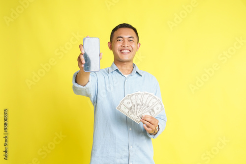 Asian man in light blue denim shirt holding blank screen phone and money banknotes dollar isolated on yellow background