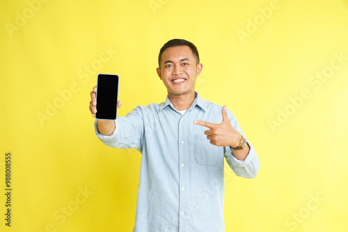Asian man in light blue denim shirt holding blank screen phone and recommended gesture isolated on yellow background